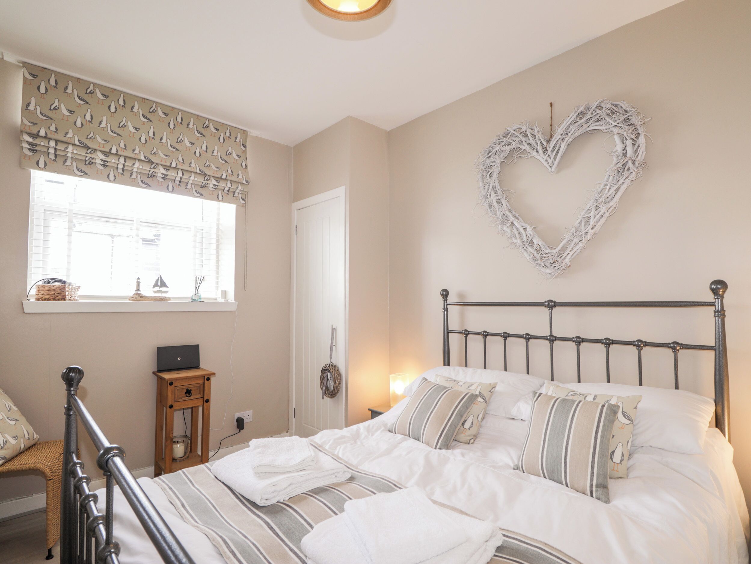 A neatly made bed with striped bedding, a heart-shaped wall decoration, a small bedside table, and a wicker chair.