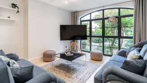 A modern living room with a large curved window, a flat-screen TV, grey sofas, and a coffee table. - SUPERB 3 BEDROOM AND OFFICE COVENT GARDEN HOUSE (London)