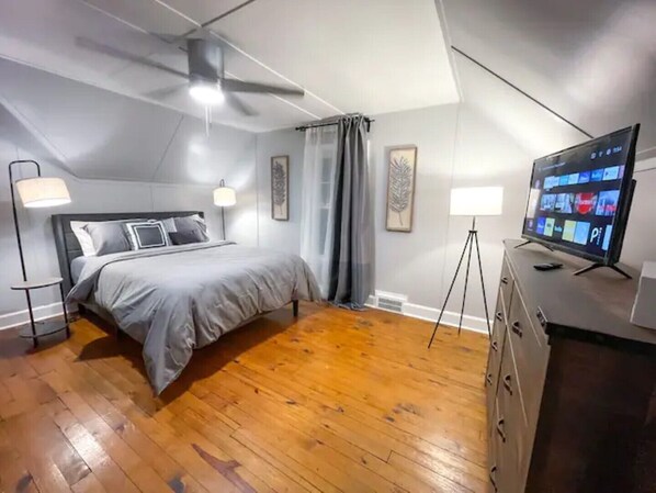 A bedroom with a bed, a ceiling fan, a TV, and a desk with a lamp.