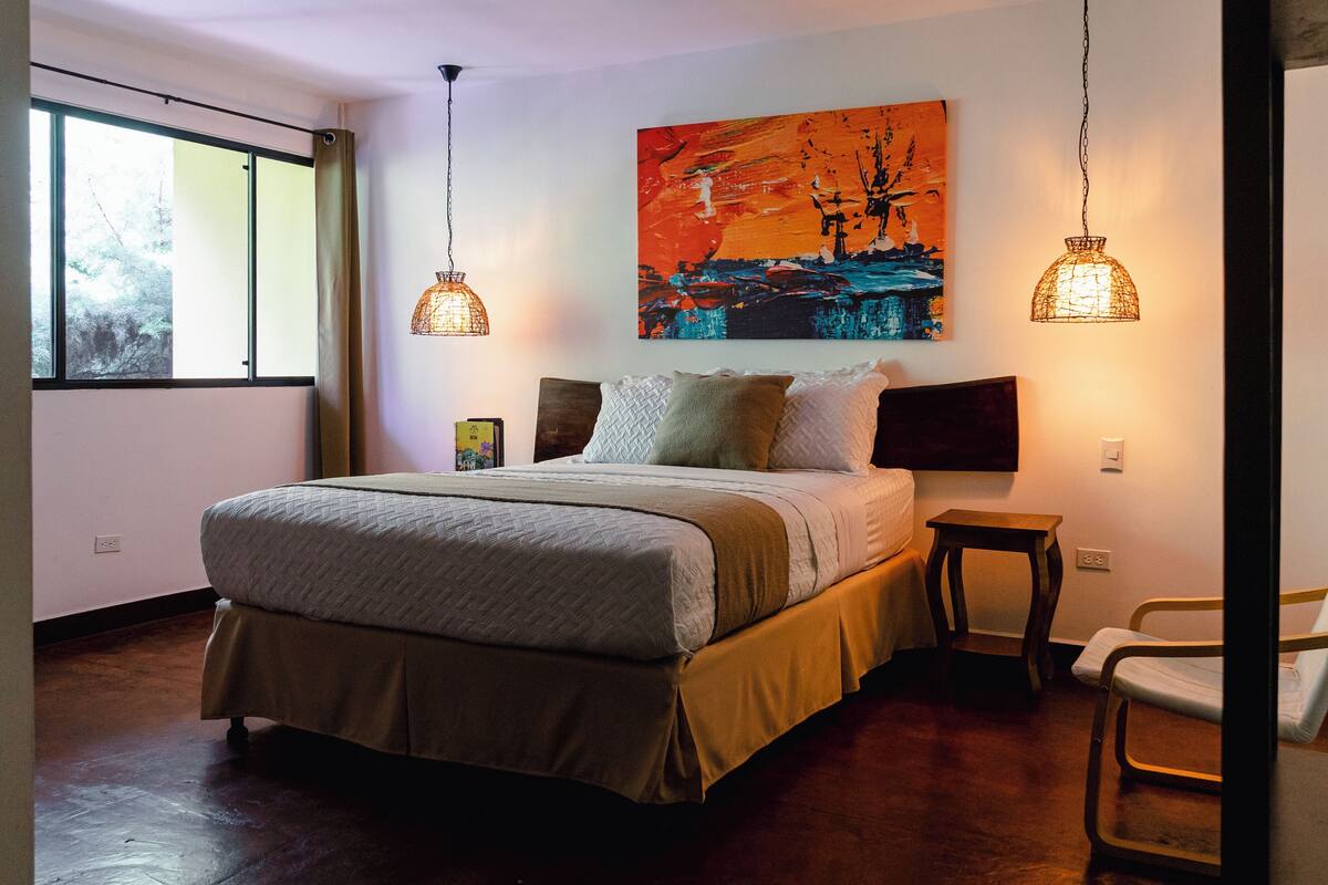 Quarto com uma cama grande, duas luminárias pendentes, um quadro na parede e uma janela com cortinas.