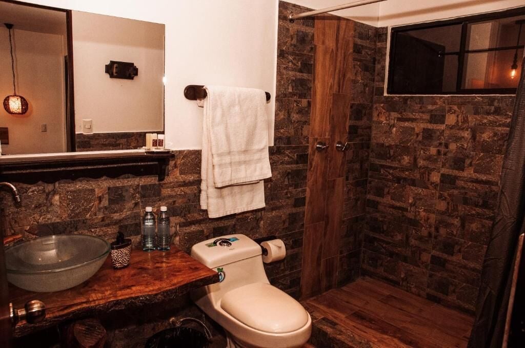 A bathroom with a stone wall, a wooden vanity, a glass bowl sink, a toilet, and a towel hanging on the wall.