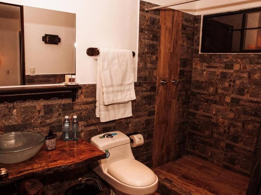 A bathroom with a stone wall, a wooden vanity, a glass bowl sink, a toilet, and a towel hanging on the wall.