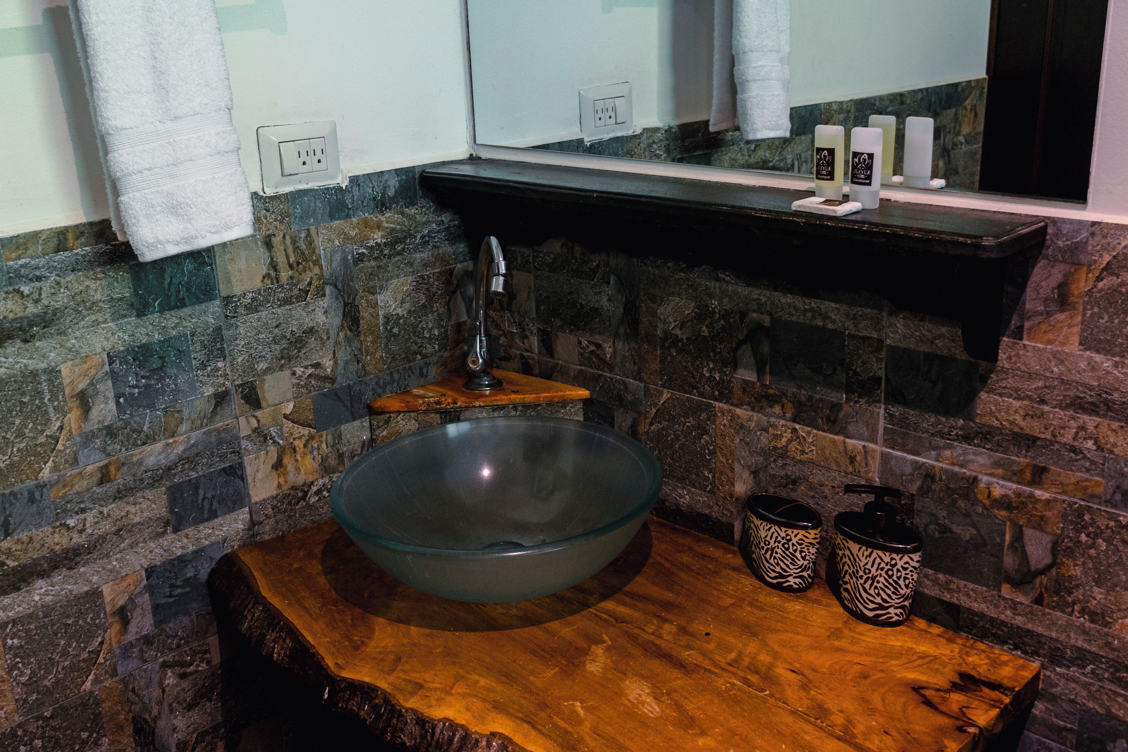 A modern bathroom with a large glass bowl sink, wooden vanity, and stone walls.