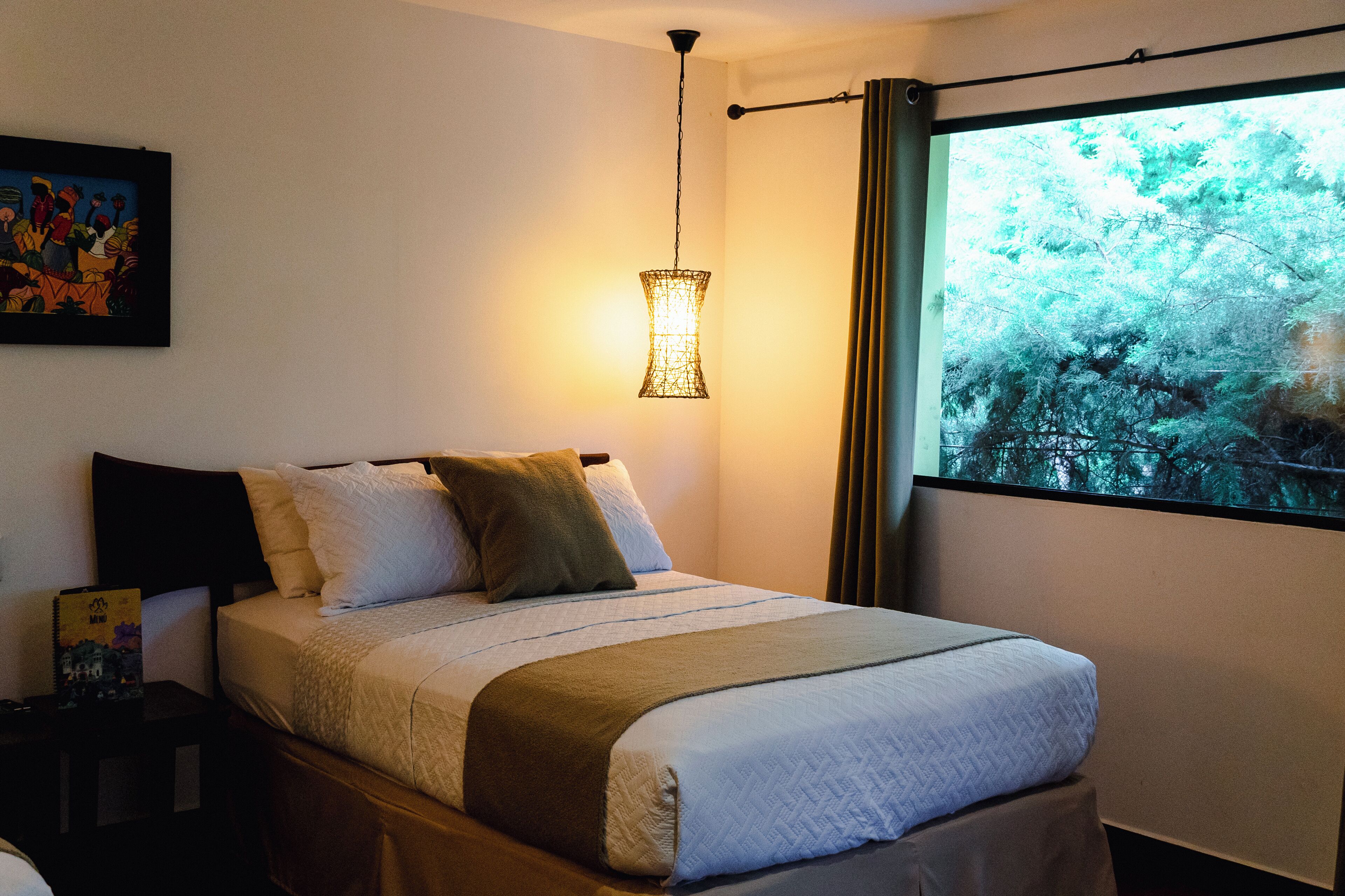 A neatly made bed with a white and brown bedspread, a hanging light fixture, and a window with a view of trees.