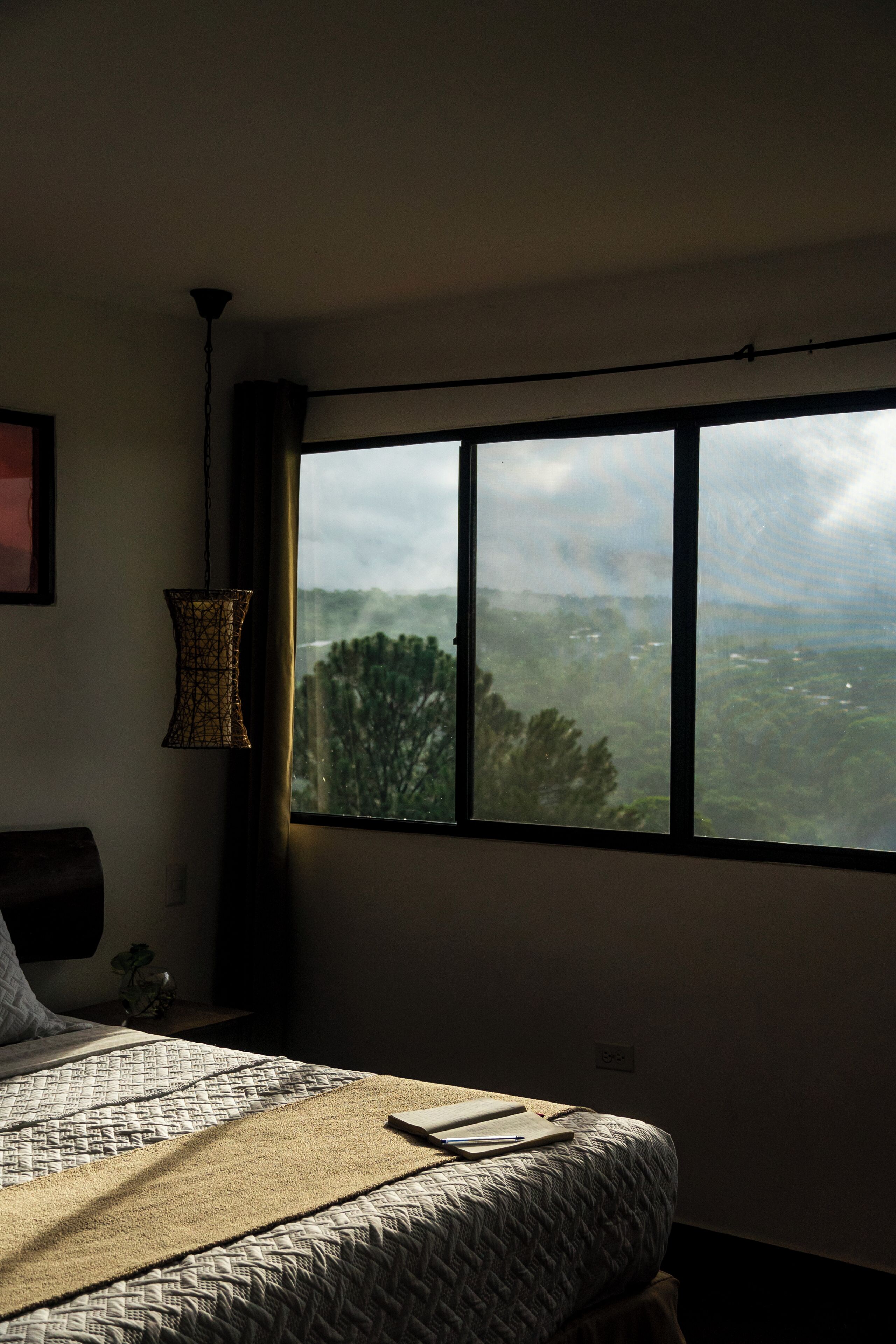 A bedroom with a large window, a bed with a patterned bedspread, a hanging lamp, and a view of a landscape.