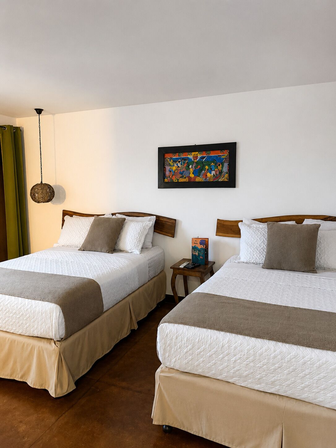 A hotel room with two beds, a painting on the wall, a hanging light fixture, and a small table with a book.