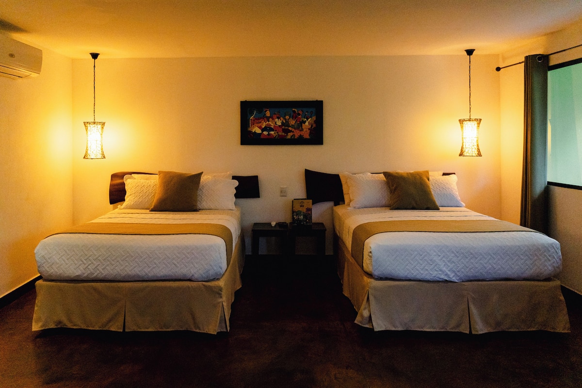 Quarto de hotel com duas camas, uma mesa de cabeceira, um quadro na parede e uma janela com cortinas.