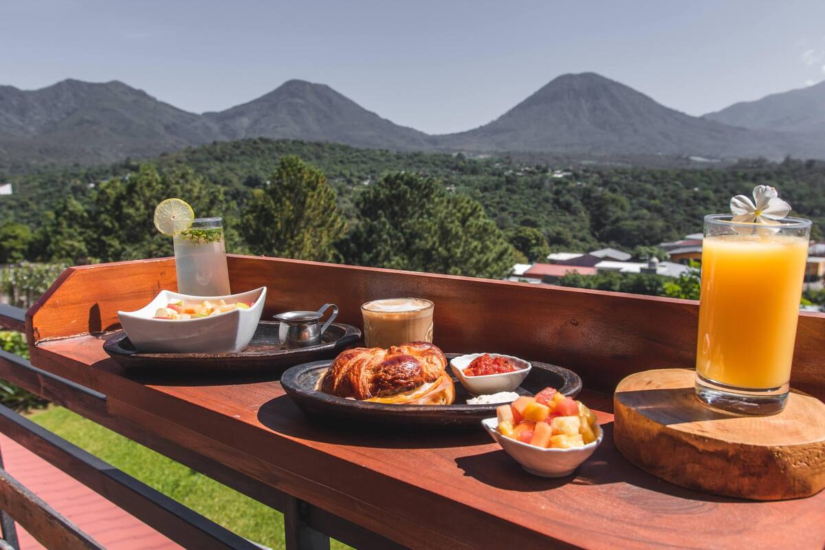 Bandeja de café da manhã com croissant, frutas, café e suco numa varanda com vista para as montanhas.