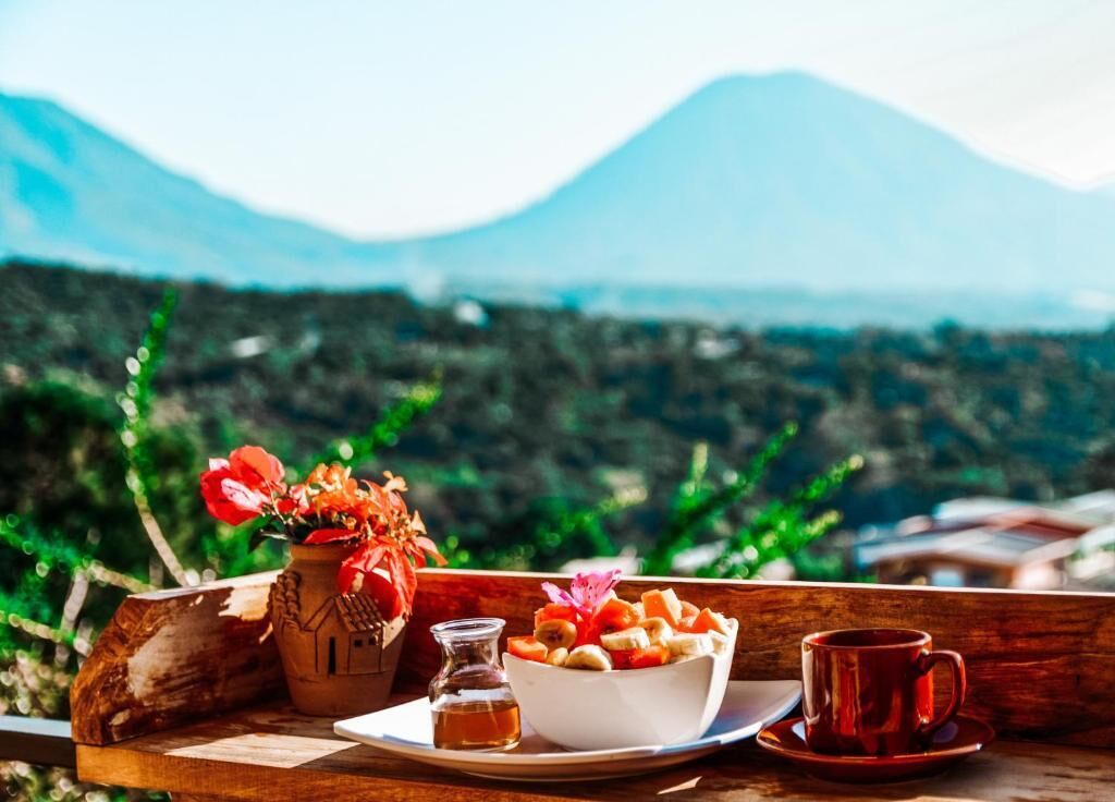 A wooden table with a bowl of fruit, a small bottle, and a red cup, set against a mountainous backdrop.