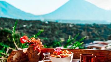 A wooden table with a bowl of fruit, a small bottle, and a red cup, set against a mountainous backdrop.