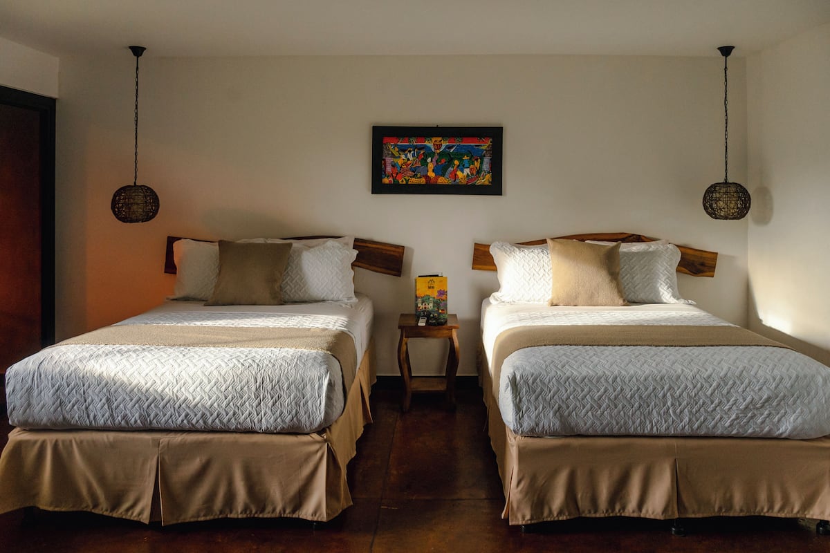 Quarto de hotel com duas camas, uma mesa pequena e uma obra de arte colorida na parede.