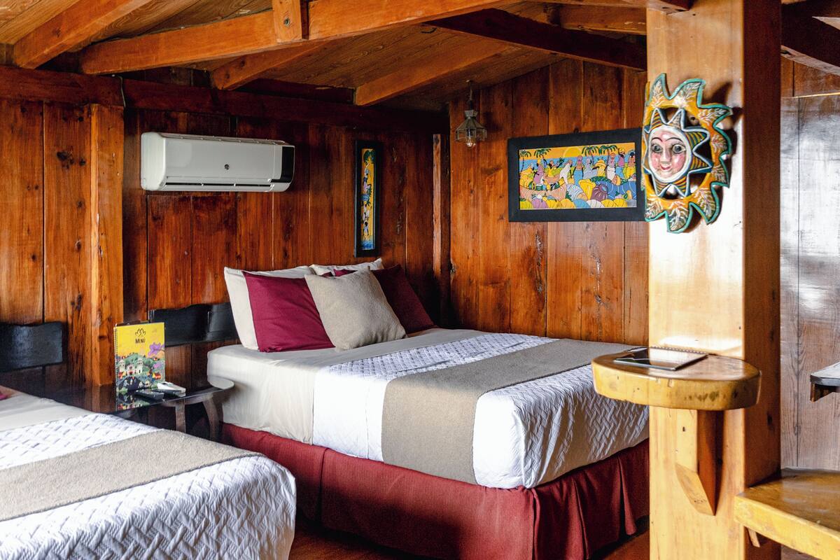 Quarto tipo cabana de madeira com duas camas, teto de madeira, ar condicionado fixado na parede e uma pintura colorida.