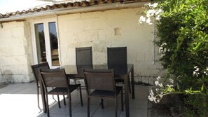 An outdoor patio with a table and four chairs, a tiled floor, and a white wall with a window and door.