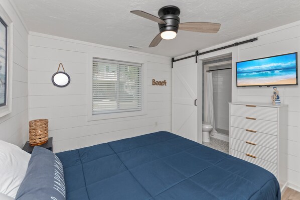 A bedroom with a bed, a ceiling fan, a TV, a window with blinds, a wall-mounted mirror, and a wardrobe.