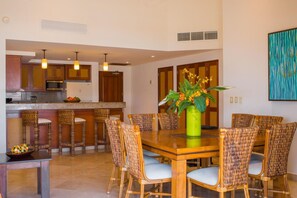 A modern kitchen with wooden cabinets, a microwave, and a refrigerator. There is a dining area with a wooden table and wicker chairs. A vase with flowers is on the table. - Luxurious Penthouse Suite on the Ridge or Beachfront side- Playa Grande Resort (Cabo San Lucas)