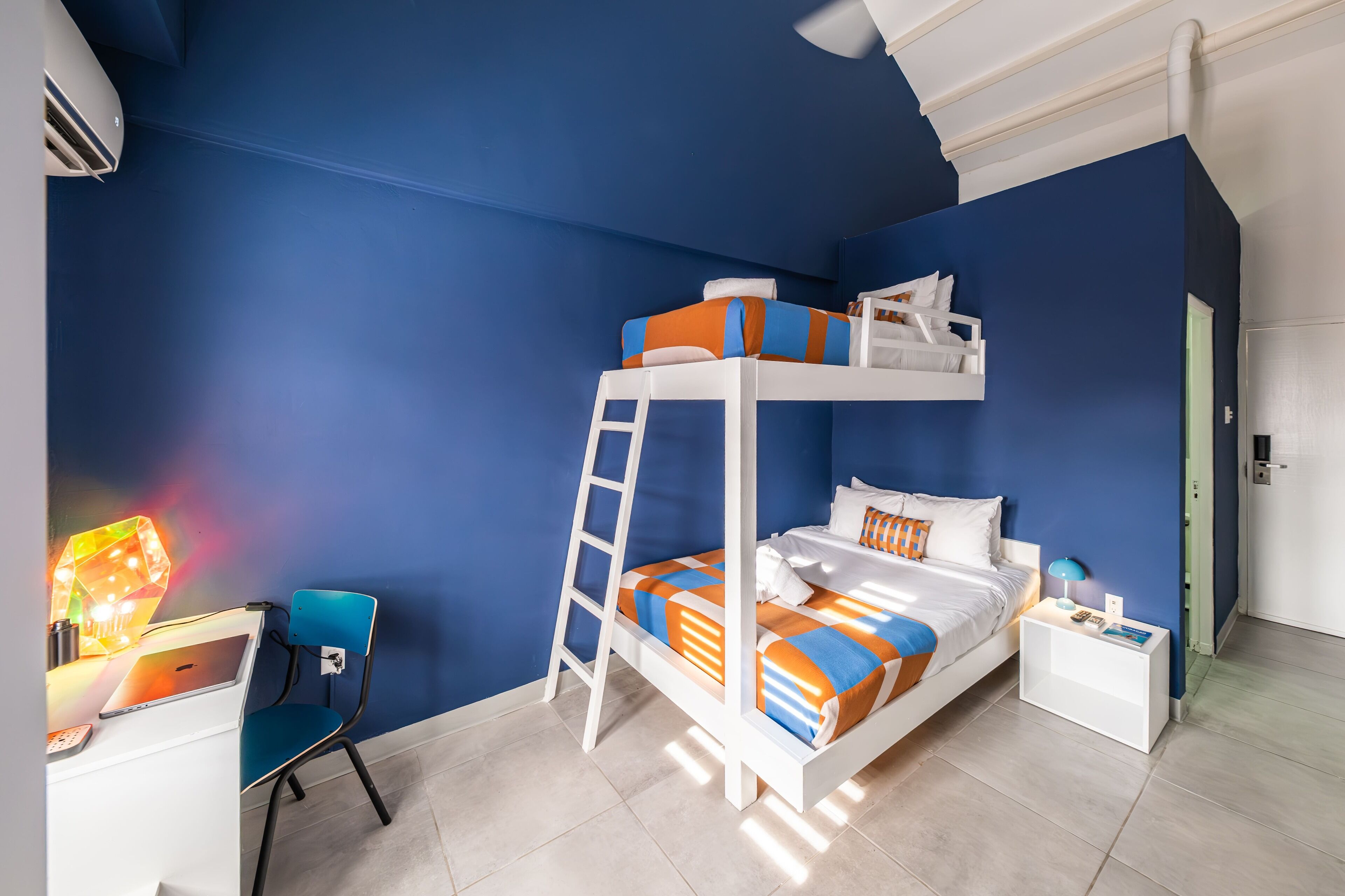 A bunk bed with a ladder, a desk with a laptop, a chair, and a bedside table with a lamp.