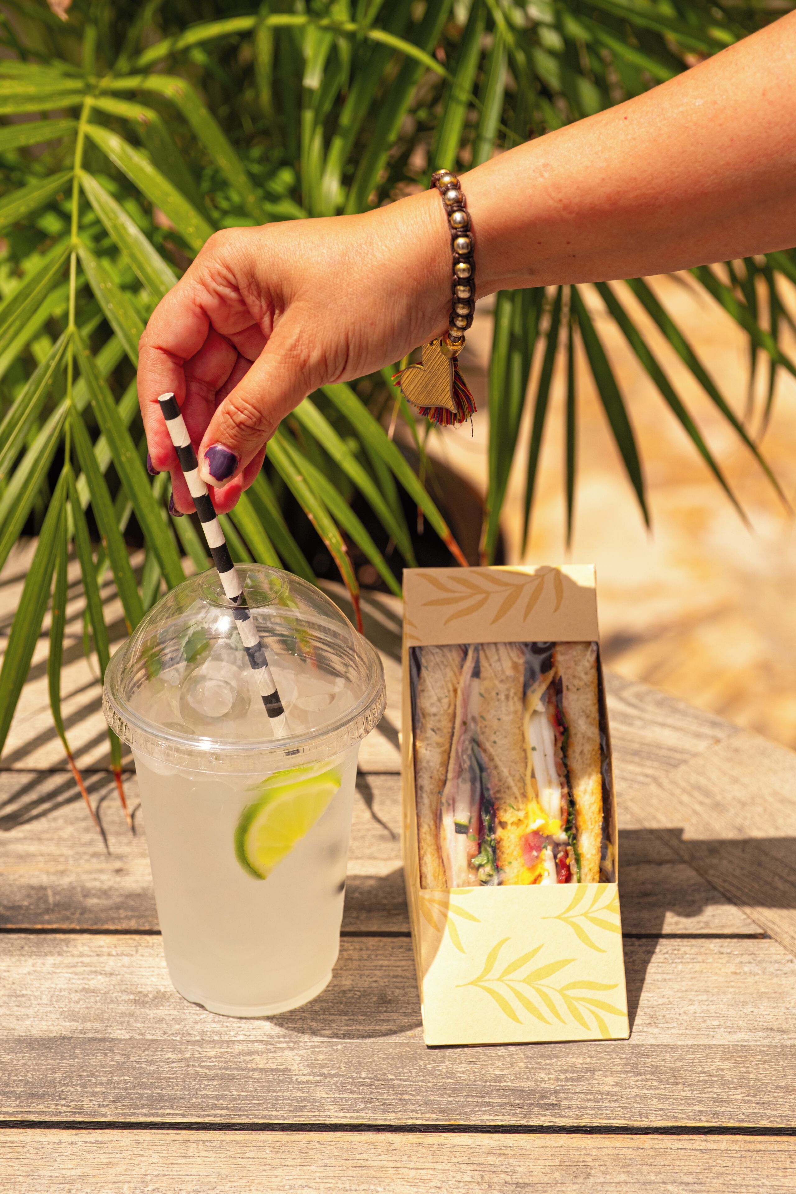 Une main tenant une paille rayée, un gobelet en plastique transparent avec une tranche de citron vert, et un sandwich dans une boîte en papier, le tout posé sur une surface en bois.