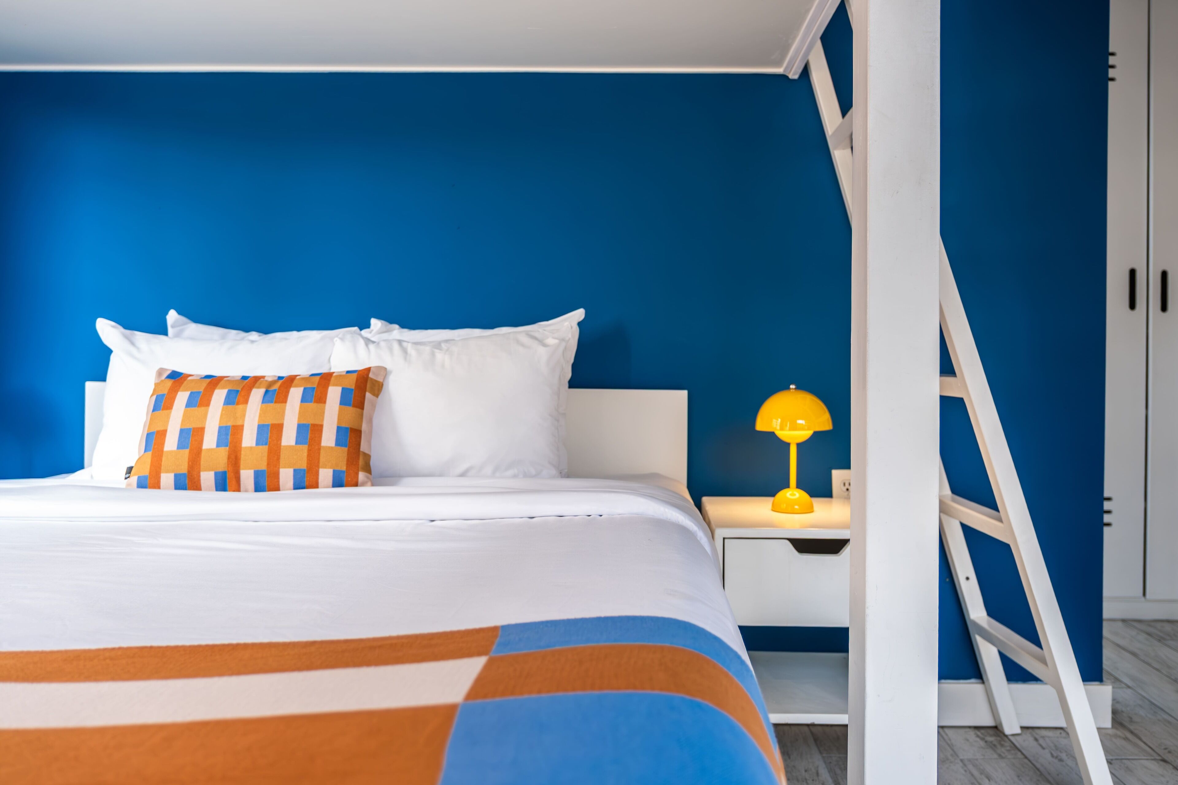 A bed with white and orange bedding, a yellow lamp on a bedside table, and a white ladder against a blue wall.