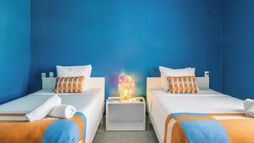 Ein Hotelzimmer mit zwei Betten, jeweils mit weißer Bettdecke und orange-blauen Kissen. Zwischen den Betten befindet sich ein Nachttisch mit einer Lampe.
