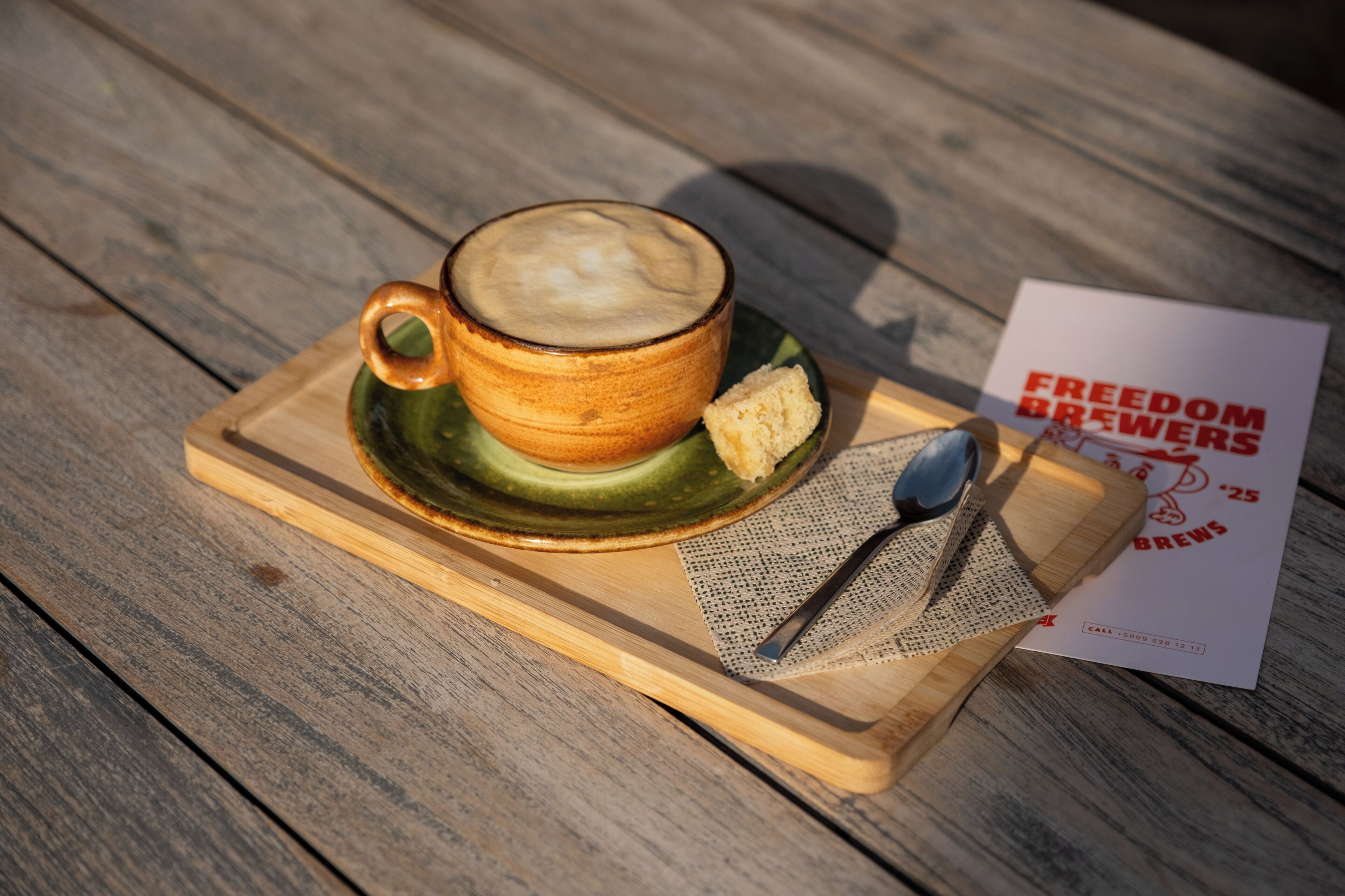 A wooden table with a ceramic cup of coffee, a biscuit, a spoon, and a menu.