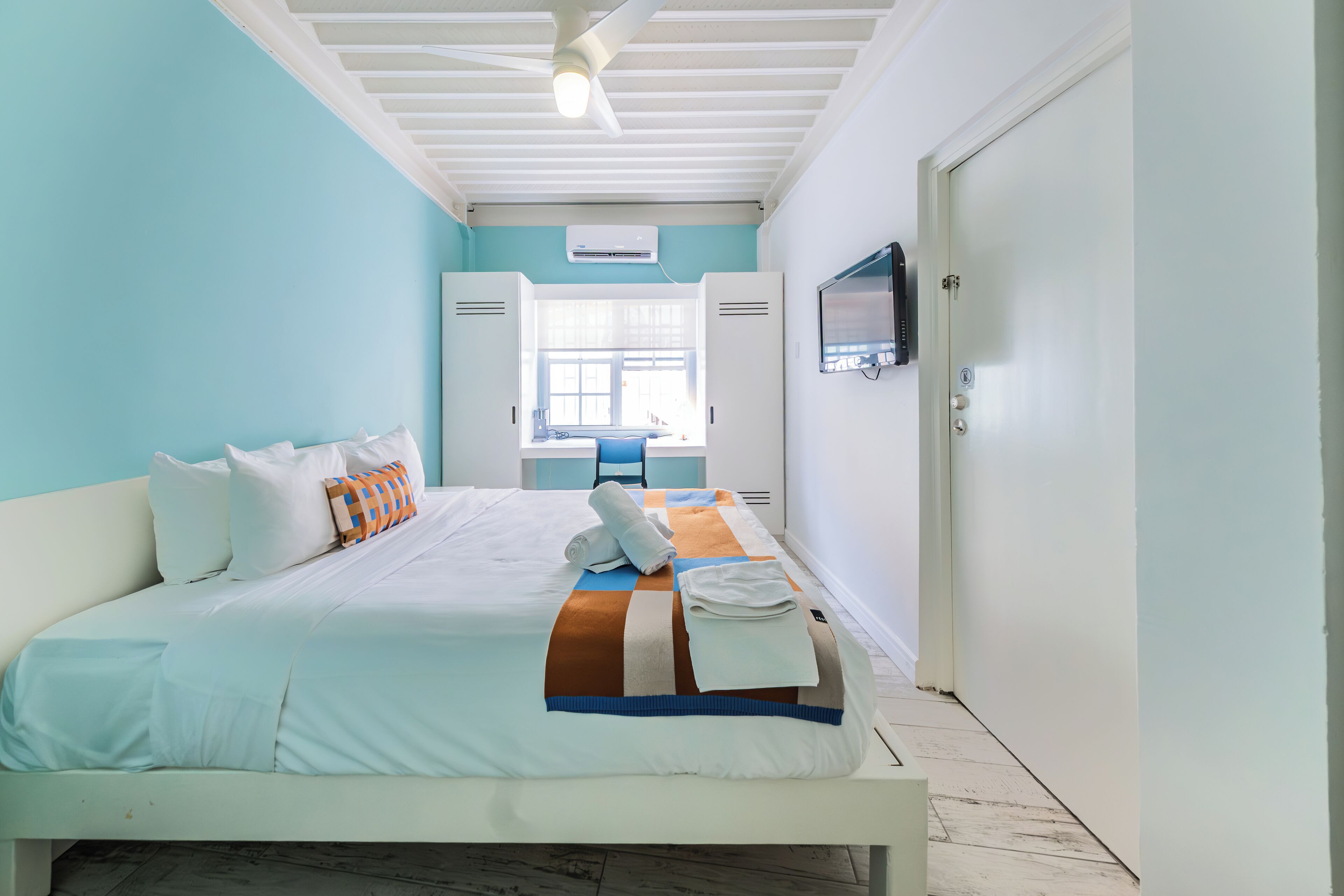 A neatly made bed with white linens and a striped blanket, a ceiling fan, and a TV mounted on the wall.
