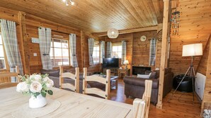 A wooden cabin interior with a dining table, chairs, a sofa, a television, and a fireplace. - Sun View Lodge (Llanbedr)