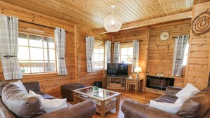 A log cabin interior with a wooden floor, leather sofas, a television, a fireplace, and plaid curtains. - Sun View Lodge (Llanbedr)