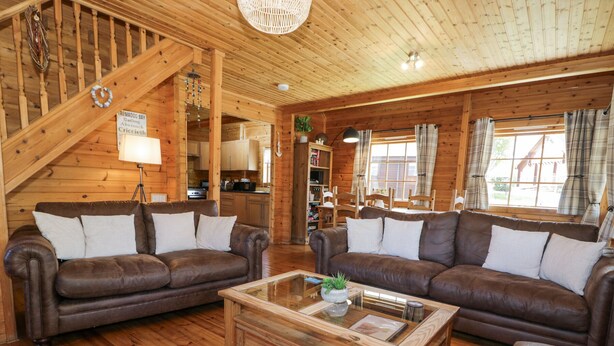 A cozy cabin interior with two leather sofas, a wooden coffee table, and a staircase leading to a loft area. - Sun View Lodge (Llanbedr)