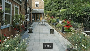 A narrow courtyard with a brick building on one side, a small table and chair in the center, and various potted plants and flowers around the perimeter.
