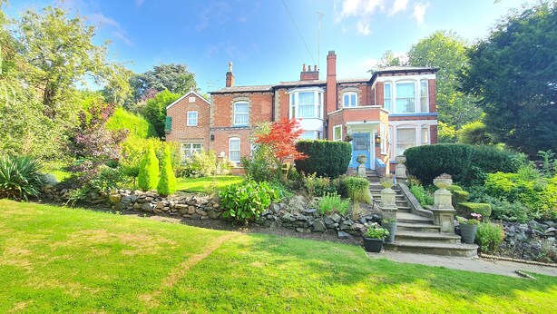 A large, two-story brick house with a well-maintained garden, stone steps leading to the front door, and a clear blue sky.