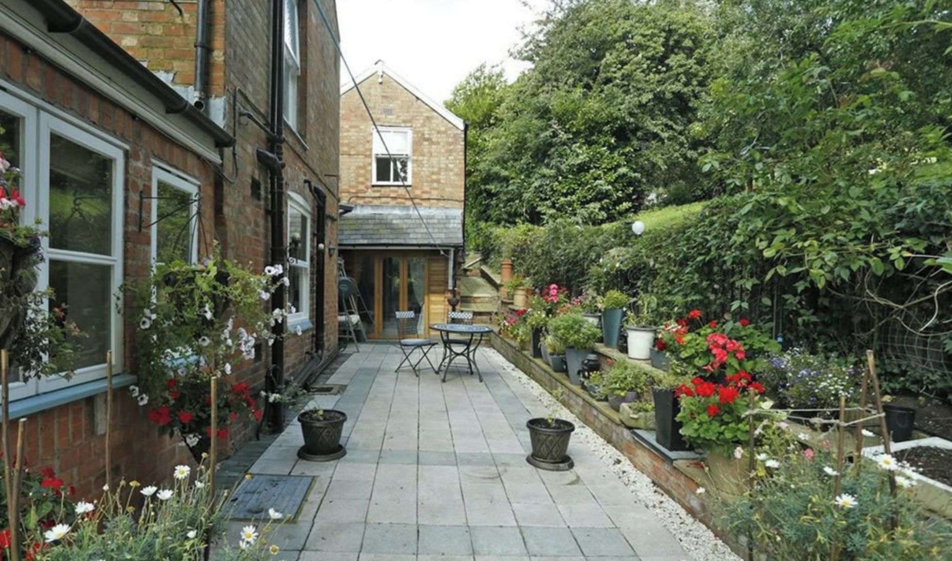 A narrow courtyard with a brick building on one side, a small table and chair in the center, and various potted plants and flowers around the perimeter.
