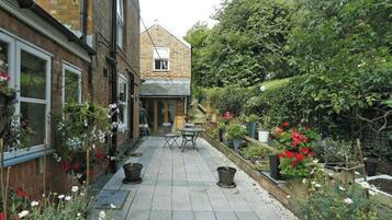 A narrow courtyard with a brick building on one side, a small table and chair in the center, and various potted plants and flowers around the perimeter.