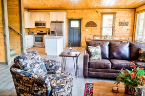 A cozy cabin interior with a kitchen, dining area, and living room furnished with a patterned armchair and a leather sofa.