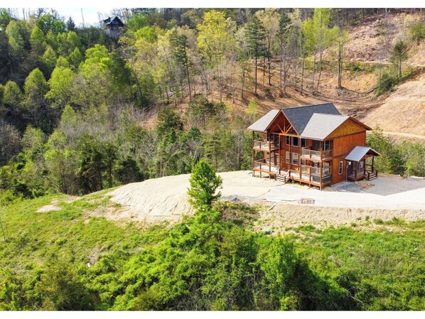A two-story log cabin with a balcony, surrounded by dense forest and a dirt path.
