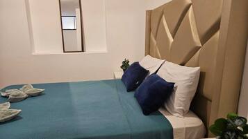 A neatly made bed with a teal bedspread and two pillows, a small bedside table with a plant, and a mirror on the wall.
