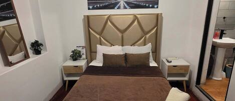 A hotel room with a large bed, a headboard with a geometric design, a nightstand with a phone, a small table with a plant, and a wall-mounted picture of an airplane.