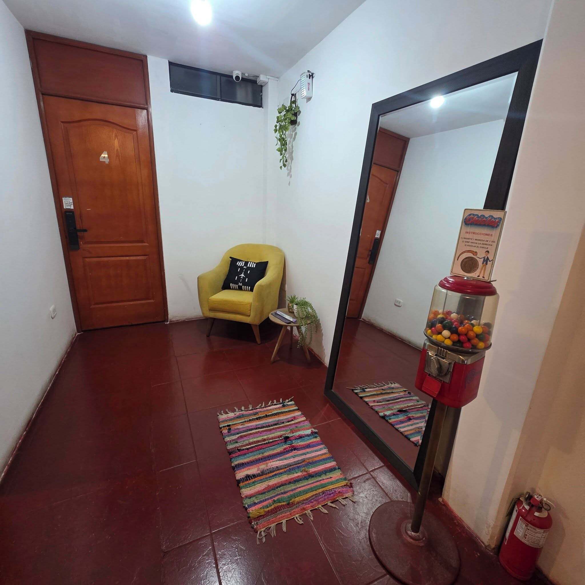 A hallway with a wooden door, a yellow armchair, a mirror, a gumball machine, and a fire extinguisher.