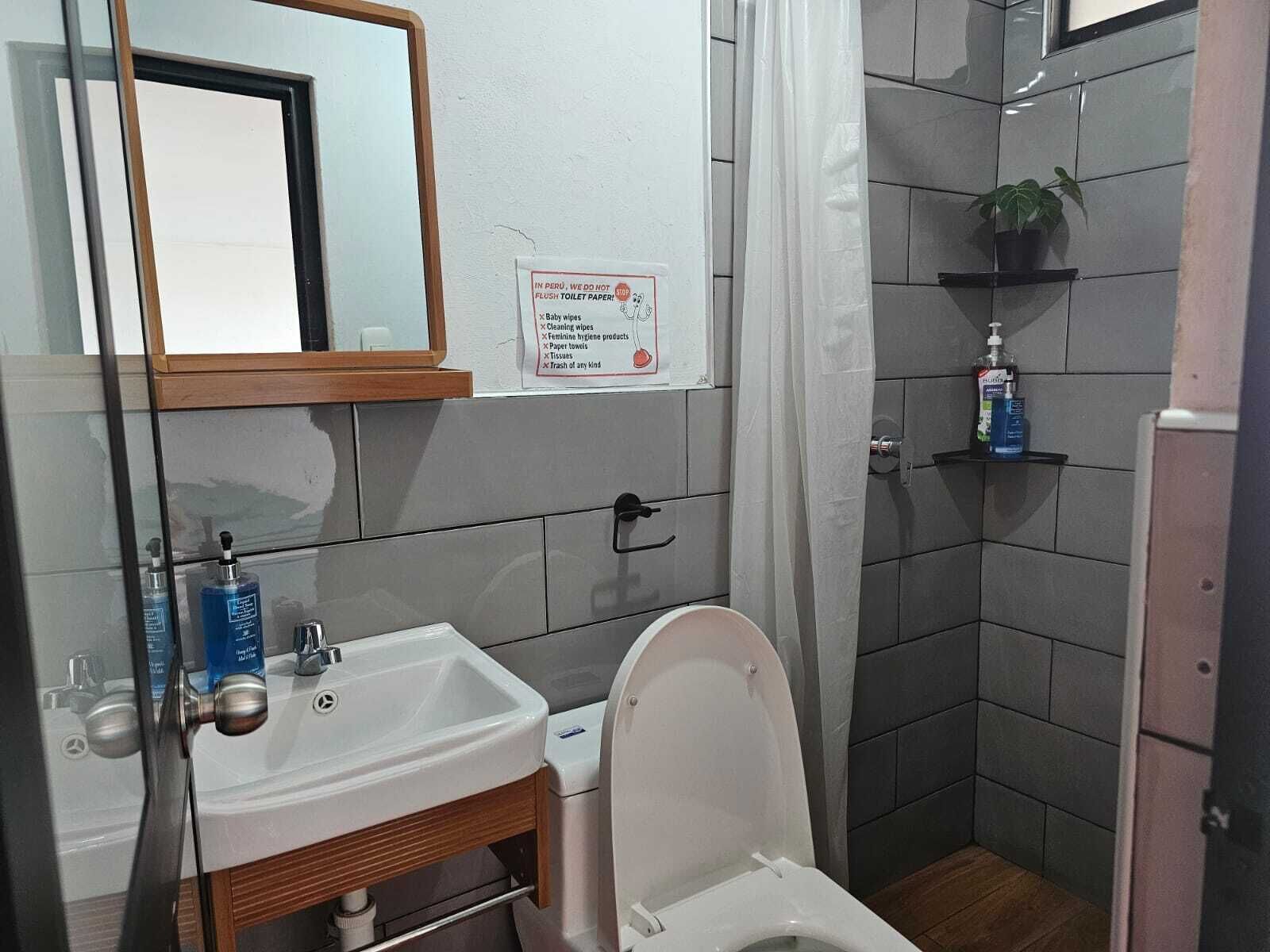 Een badkamer met een witte wastafel, een toilet, een spiegel en een douchegedeelte. Er hangt een bord op de muur met daarop een overzicht van de voorzieningen in de badkamer.