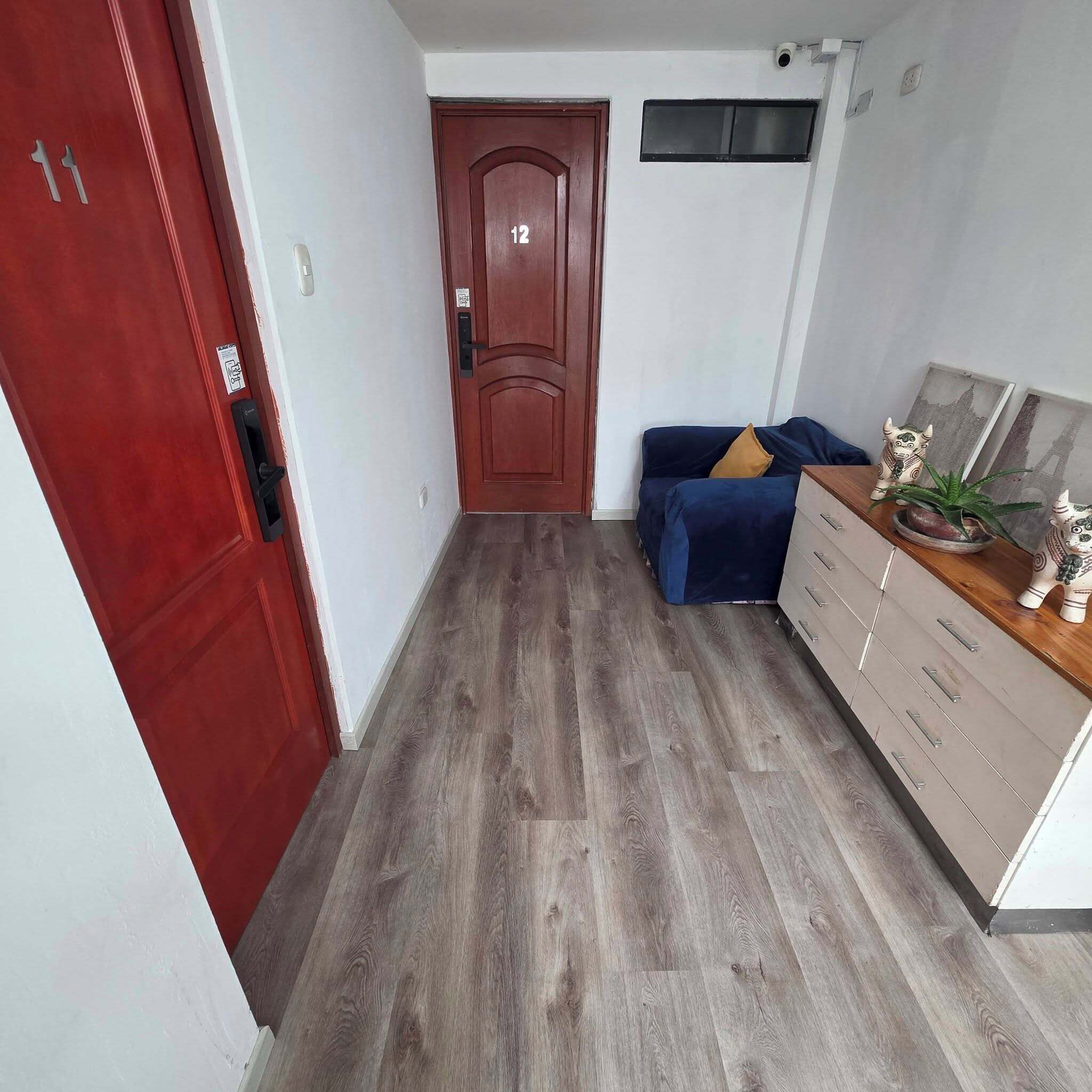 Un pasillo con piso de madera, una puerta roja con el número 12, un sofá azul con un cojín amarillo y un aparador de madera con cajones.