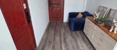 Un pasillo con piso de madera, una puerta roja con el número 12, un sofá azul con un cojín amarillo y un aparador de madera con cajones.