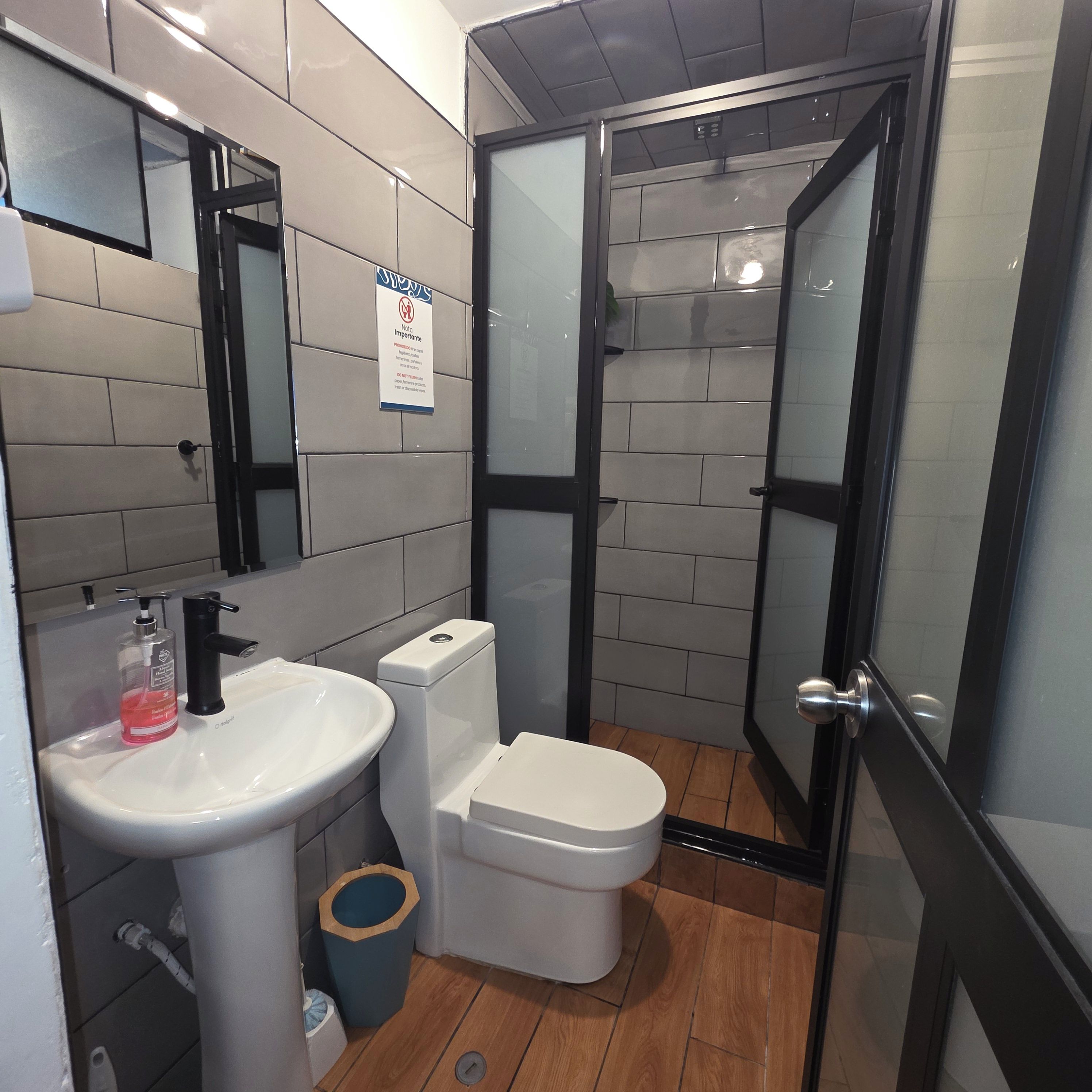 Un baño moderno con un lavamanos blanco, grifo negro y un inodoro. Hay un cubo de basura y un espejo en la pared.