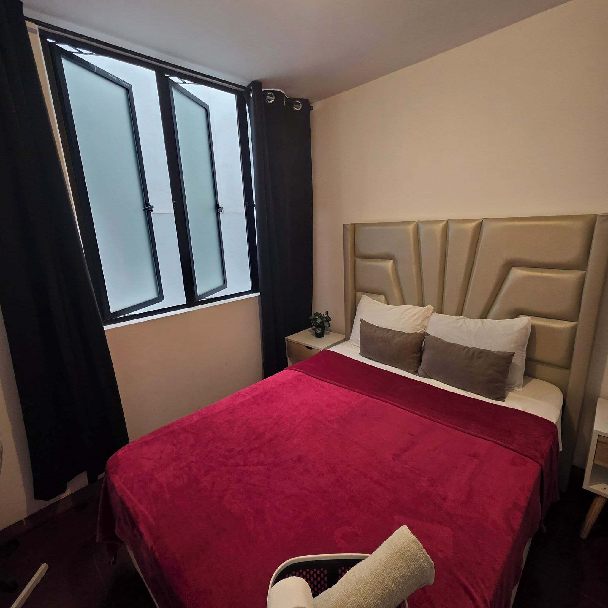 A hotel room with a bed, a nightstand, a window with curtains, and a view of the outside.