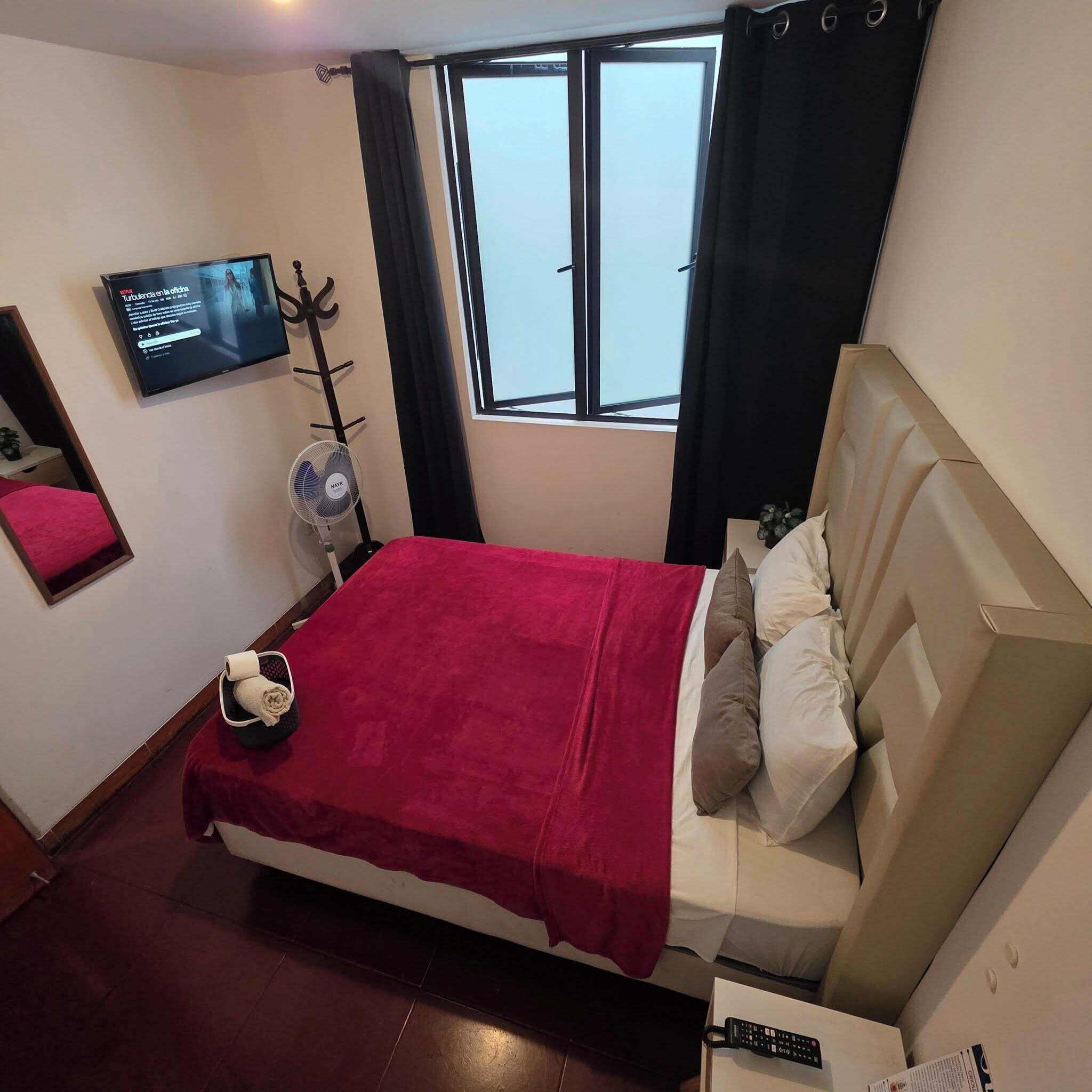 Habitación de hotel con una cama, un televisor montado en la pared, una ventana con cortinas y un ventilador.