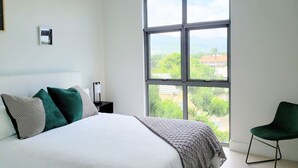 A bedroom with a large window, a bed, a chair, and a nightstand. - 24/7 Security, 2B/R Paradise w/Infinity Rooftop Pool & City Views 🌴🏊 (Kingston 10)