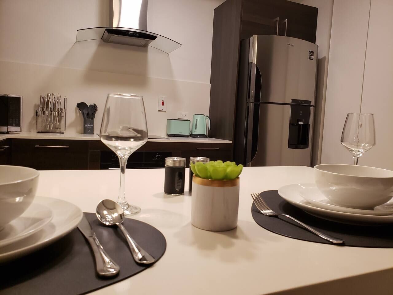 Une cuisine moderne avec une table à manger pour deux personnes, caractérisée par un design minimaliste avec un plan de travail blanc, des appareils en acier inoxydable et une plante décorative en guise de centre de table.