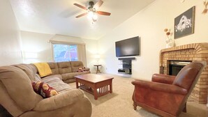 A living room with a sectional sofa, a leather armchair, a coffee table, a flat-screen TV, a fireplace, and a ceiling fan. - The Hideout-Single Level 2BR/2BA Townhouse near Highland! (Pocatello)