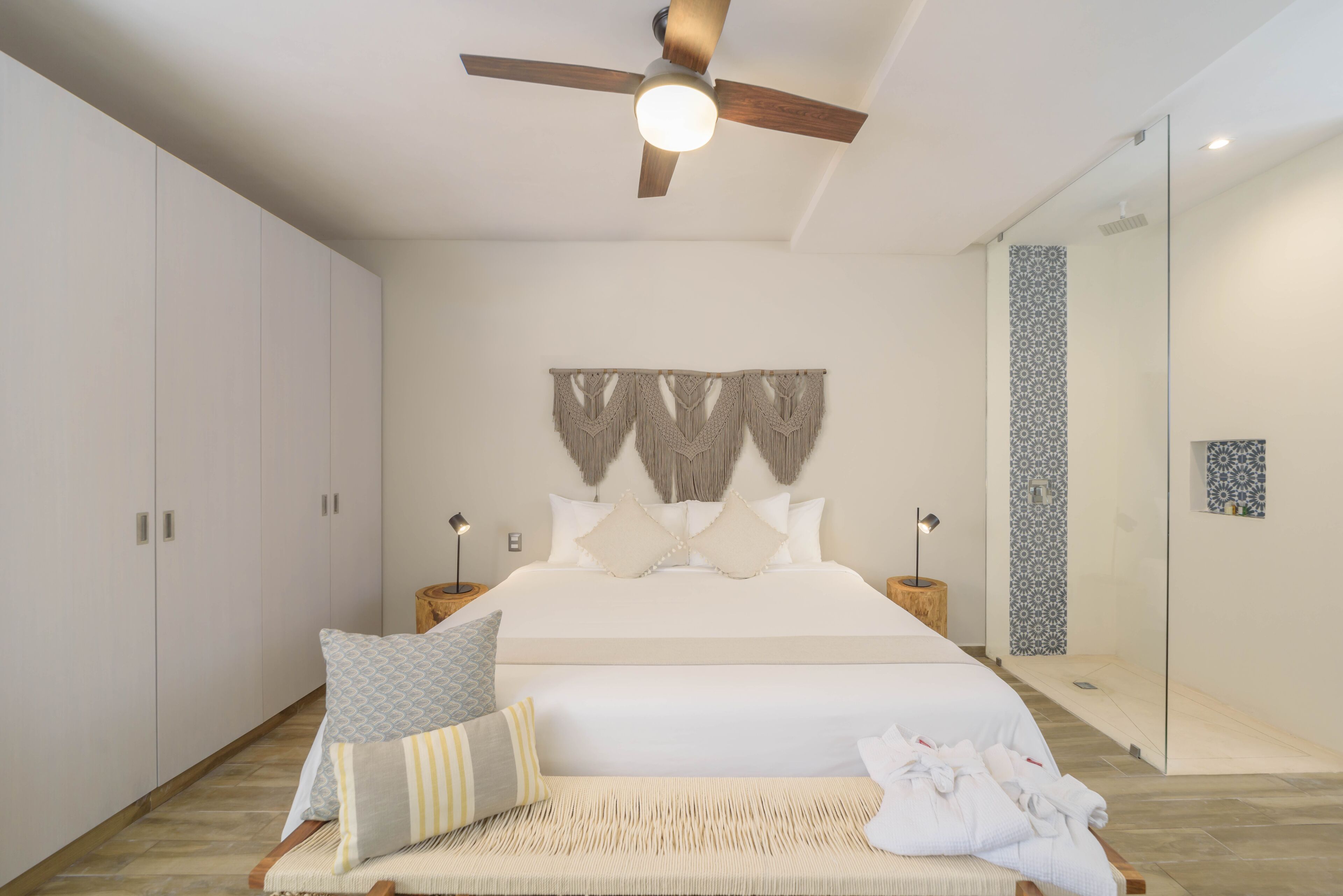 A bedroom with a bed, a ceiling fan, a bedside table with a lamp, a wall-mounted decorative piece, and a glass shower enclosure.