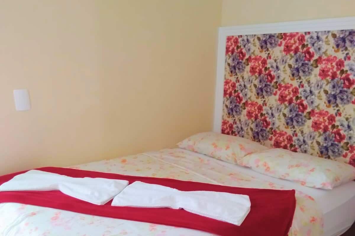 A bed with floral bedding and a red blanket, a wooden headboard with a floral pattern, and a plain wall with a light switch.