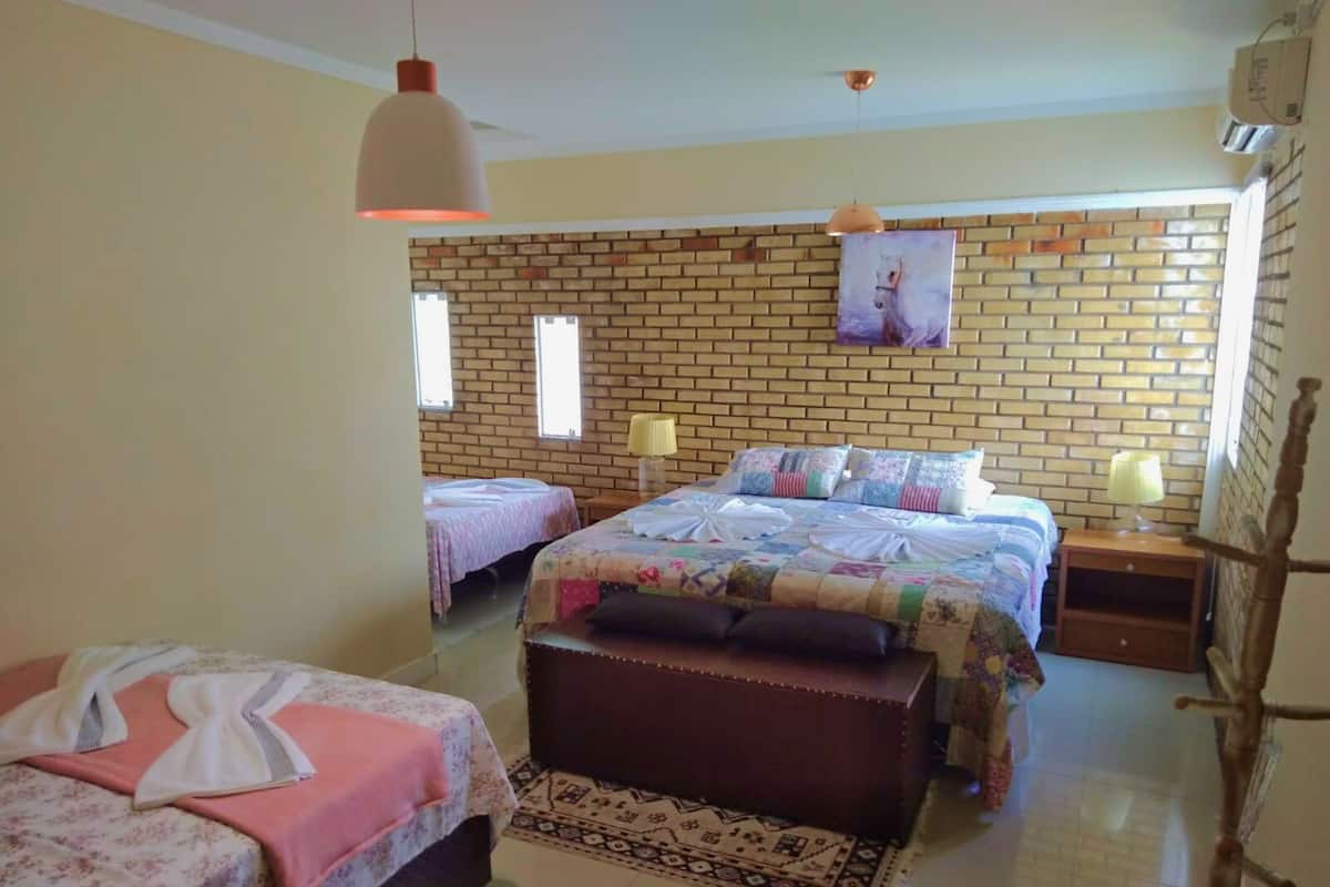 A hotel room with two single beds and a double bed, a brick wall, a window, and a hanging light fixture.