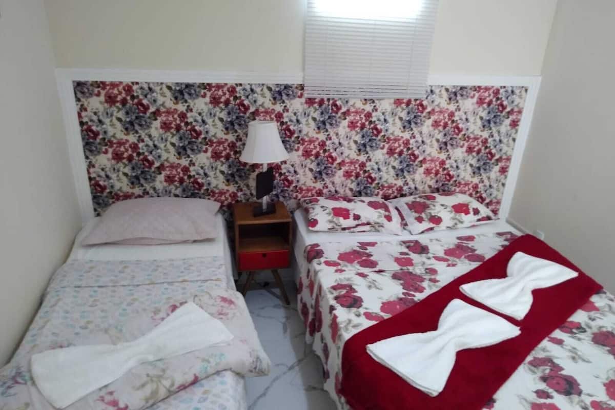 A bedroom with a floral headboard, a bedside table with a lamp, and two beds with floral bedding.